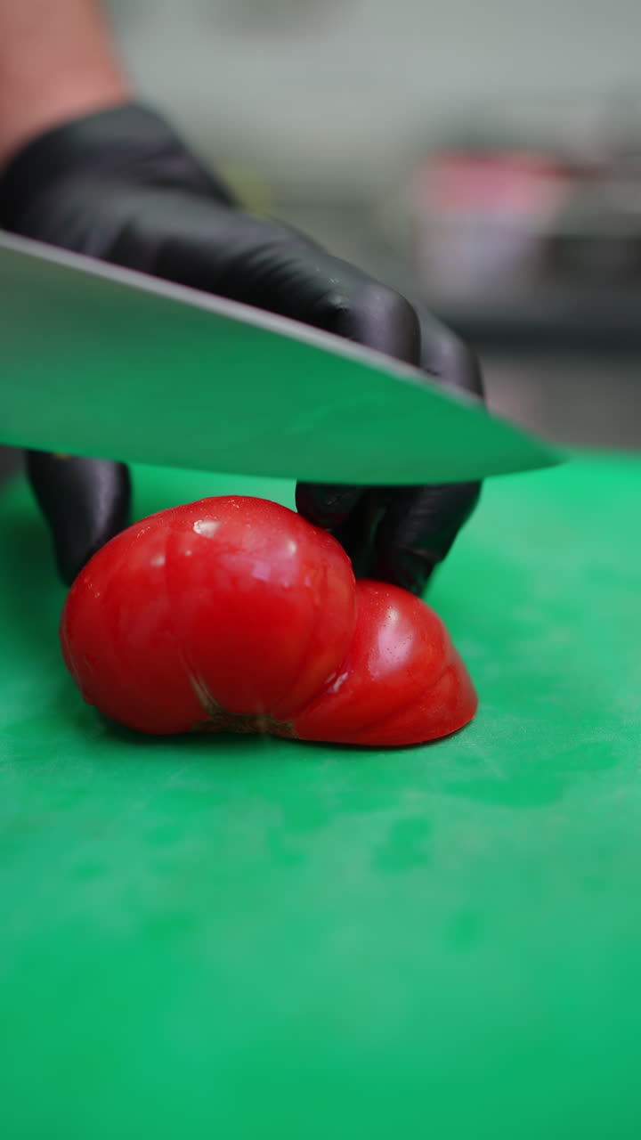 Vertical footage Sharp knife cutting fresh red tomato into uniform slices with professional precision, resting on bright green chopping surface, showcasing culinary expertise