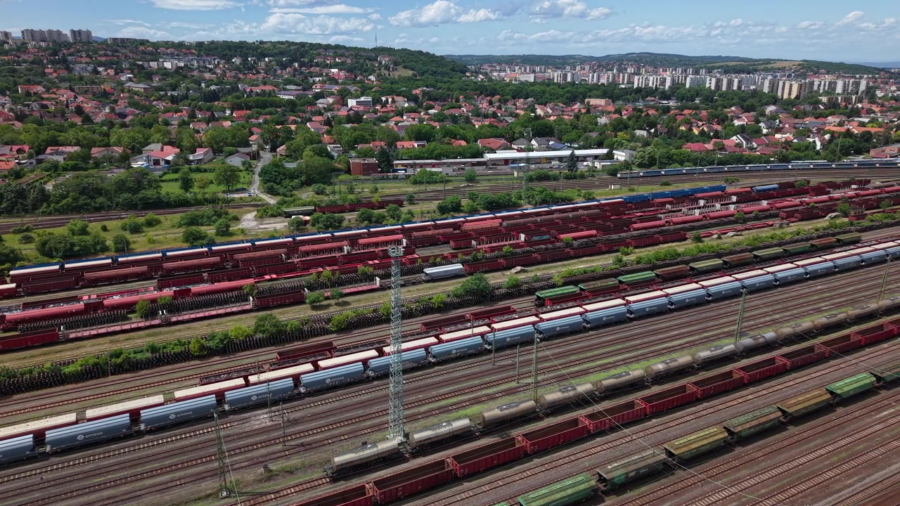 Ein ausgedehnter Rangierbahnhof zeigt zahlreiche Güterzüge und Güterwaggons auf mehreren Gleisen, vor dem Hintergrund einer Stadt und grüner Hügel.