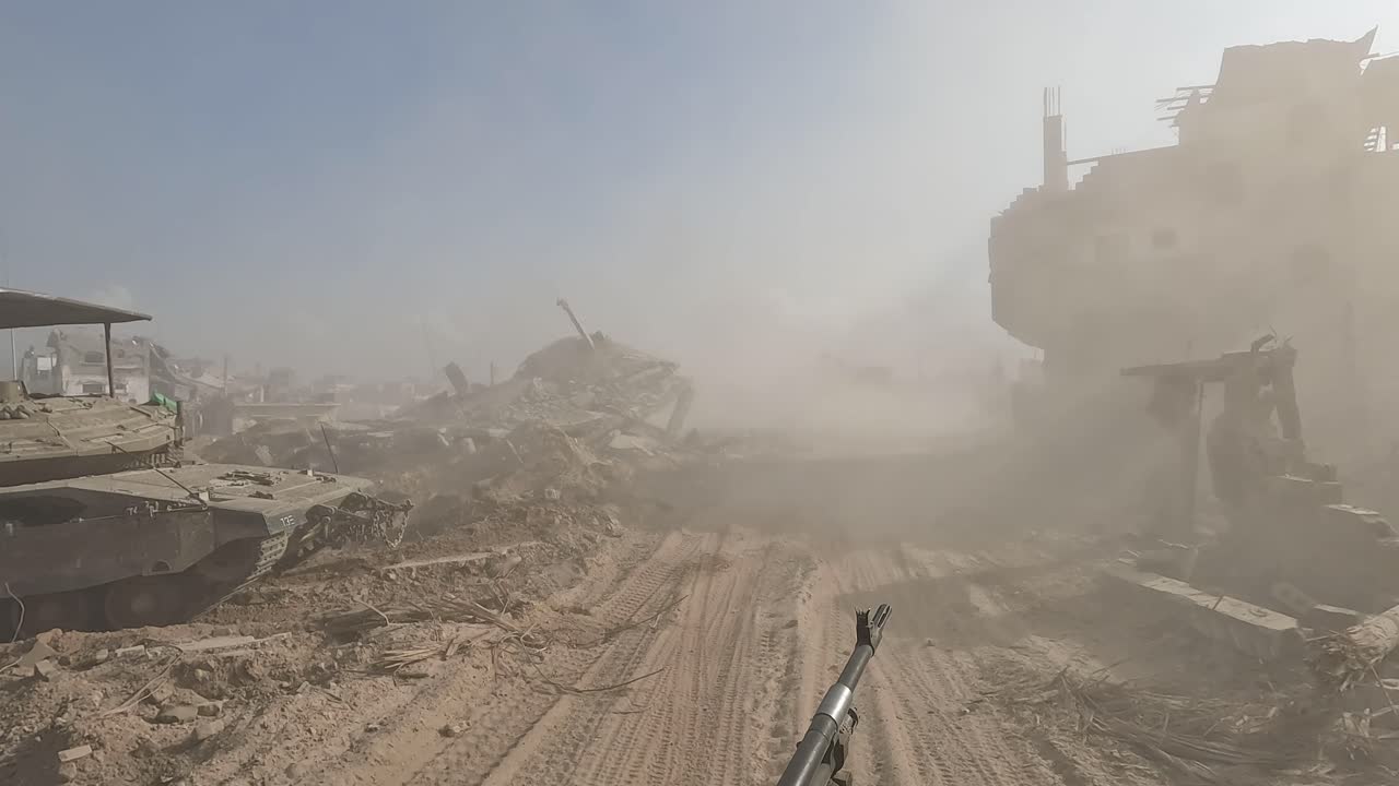 Striscia di Gaza — POV da un veicolo militare che attraversa una strada polverosa, superando un veicolo blindato e edifici gravemente danneggiati.