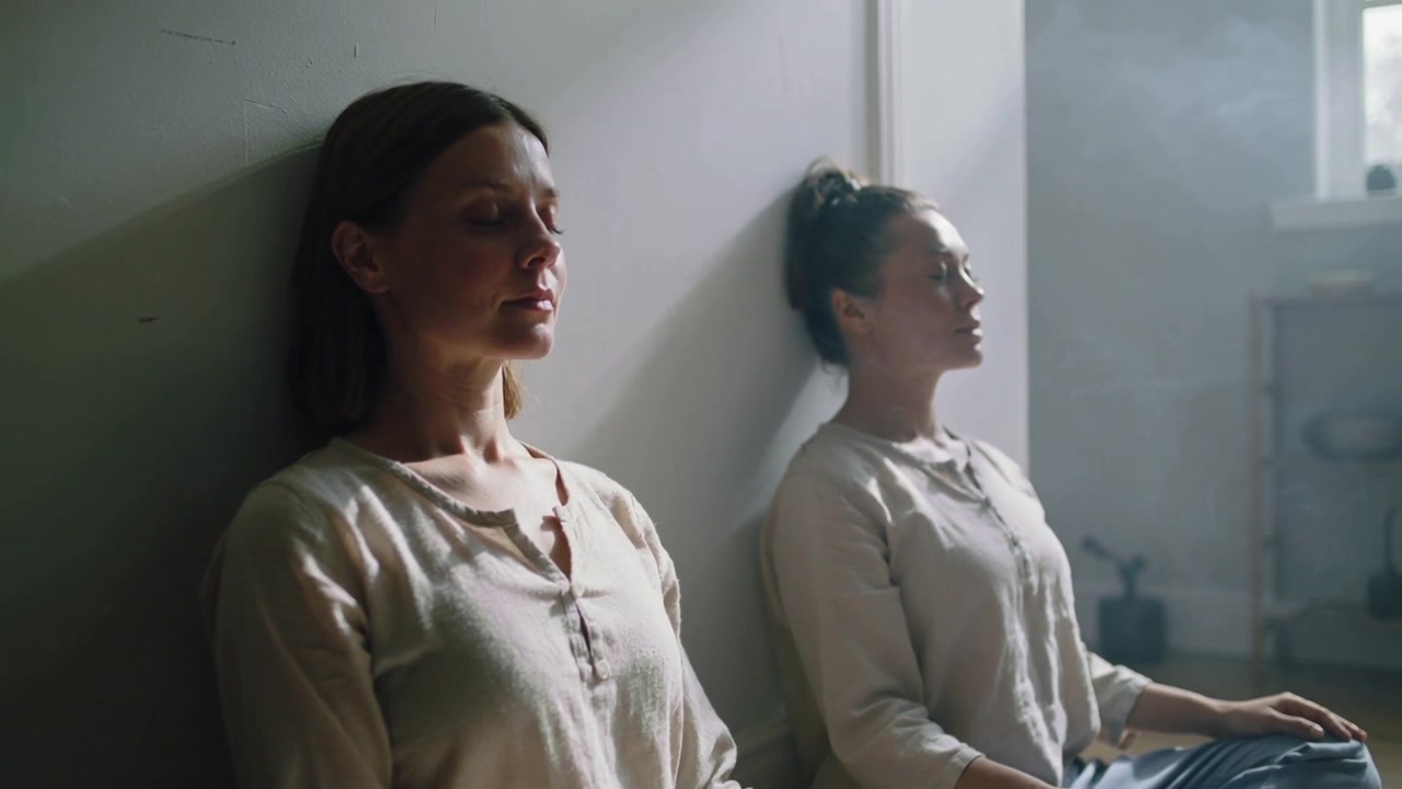 Dos mujeres sentadas en tops haciendo ejercicios de respiración para la calma en el suelo junto a la ventana, con la luz del sol entrando e incienso.