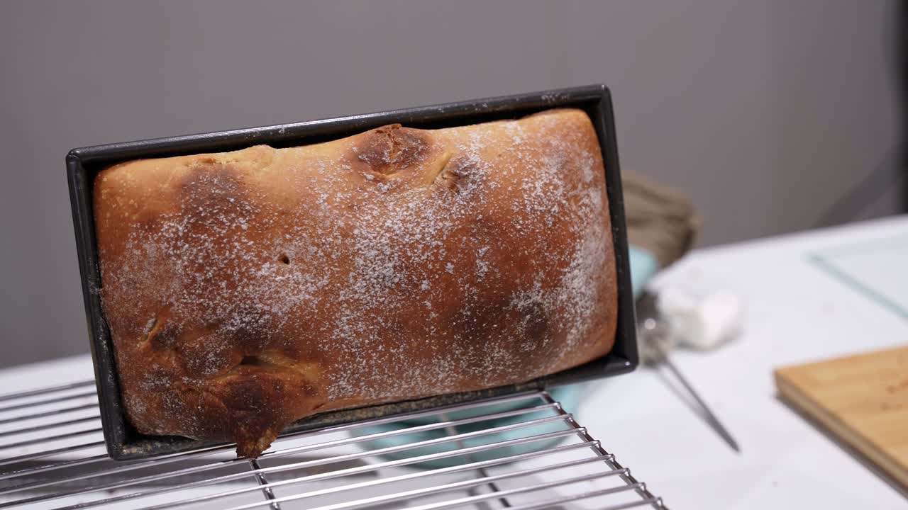 Close-up of a freshly baked homemade bread loaf dusted with flour in a rectangular metal pan, resting on a cooling rack in a kitchen setting. Artisan baking and homemade food concept