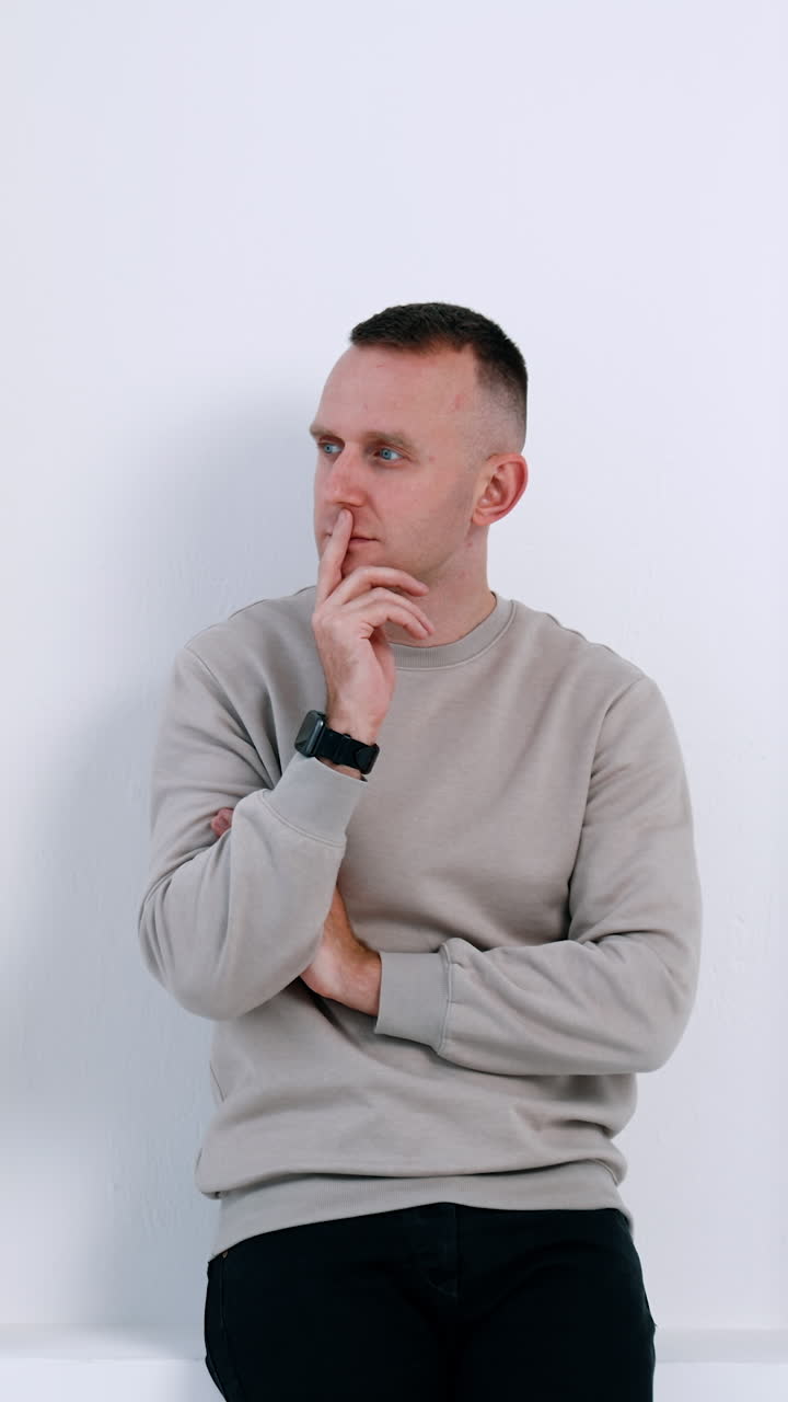 Pensive Caucasian male wearing grey sweater. Man stands at the white wall looking to the side and smiling. Vertical video