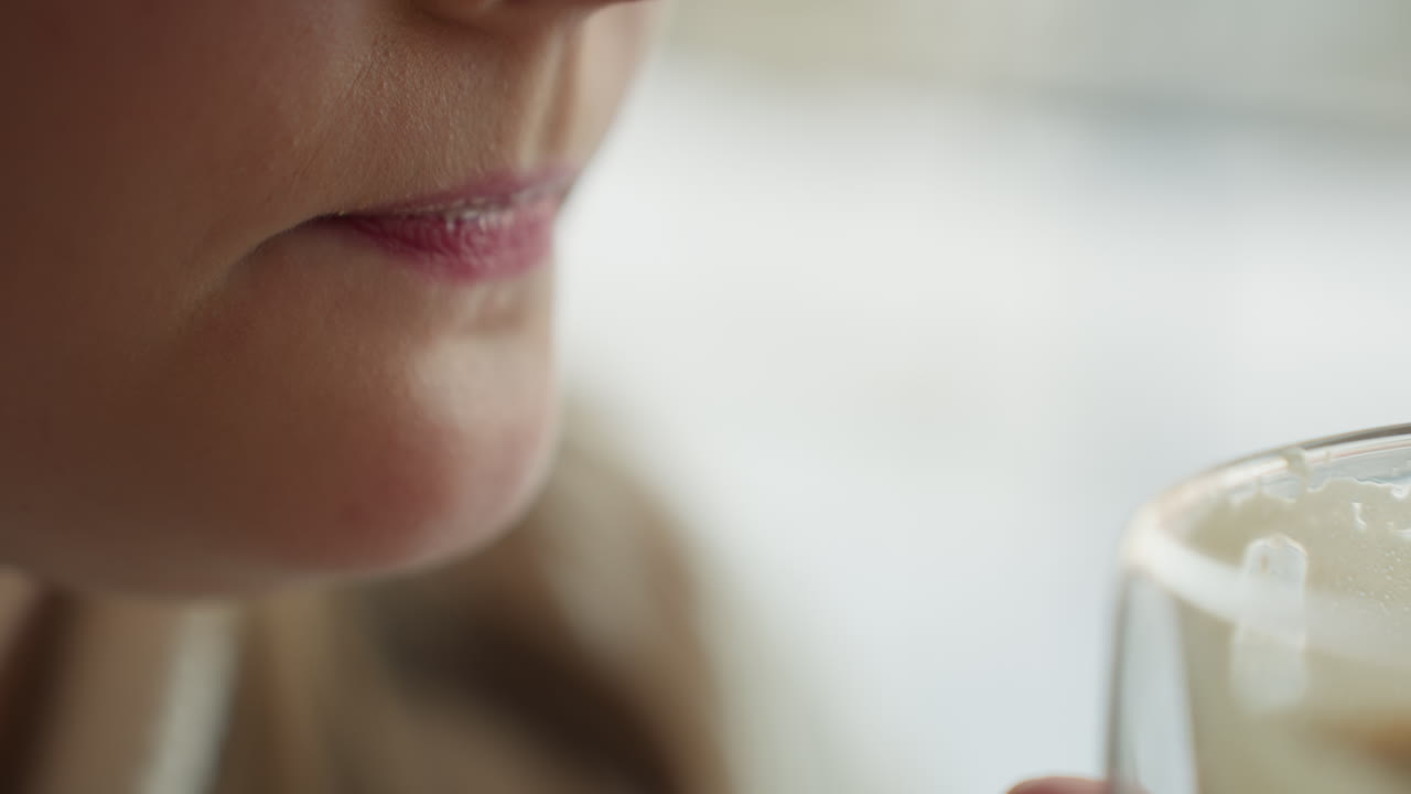 extreme close up of woman gently sipping creamy milkshake from transparent glass with both hands, soft natural light and blur in background
