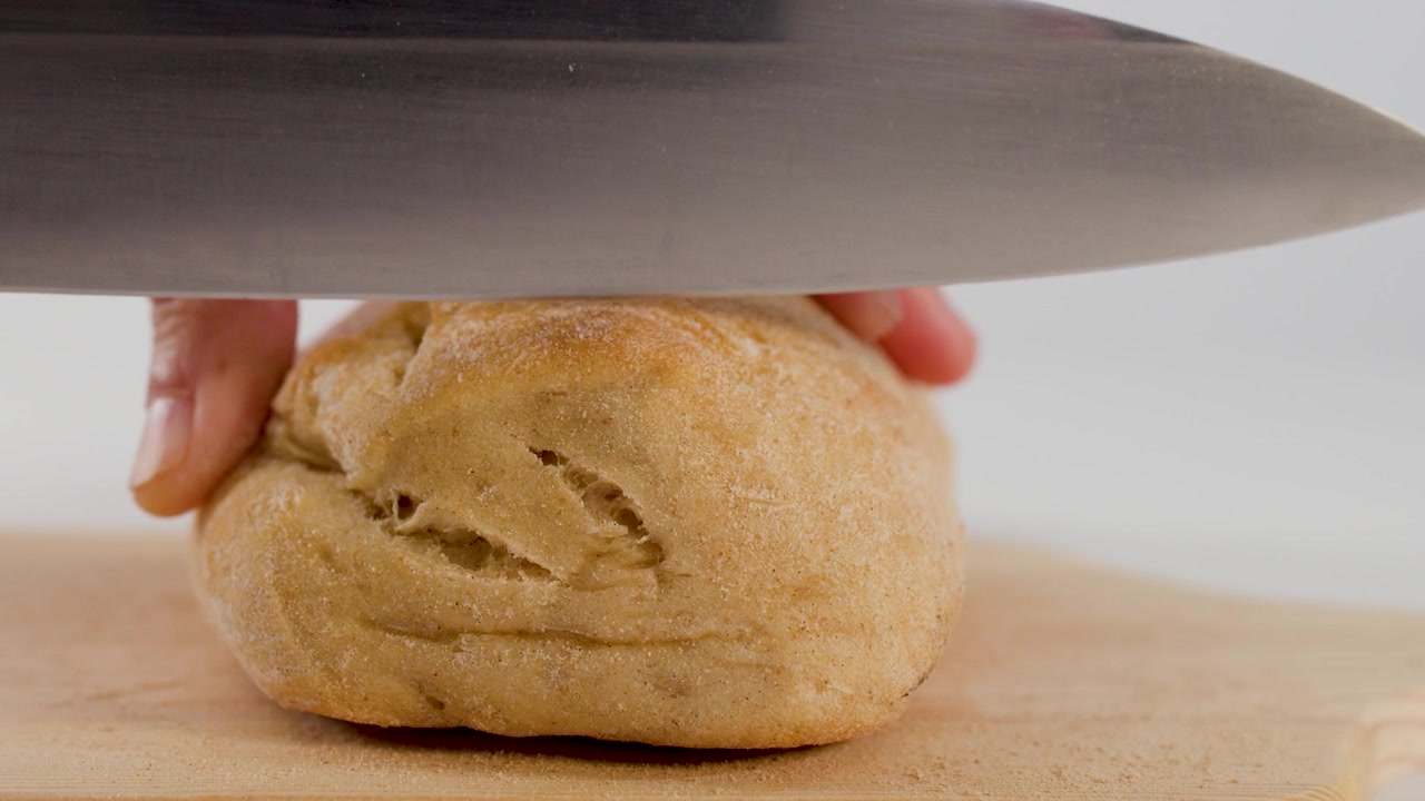 Close-up of hands slicing a crusty bread roll on a wooden board against white.