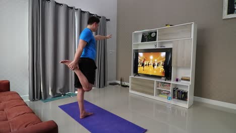 a man is stretching his legs while standing in front of a television in his living room following a fintess programme trying to burn some calories