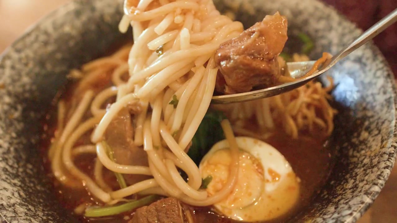 Primo piano di noodles e manzo sollevati da un brodo piccante con bacchette e un cucchiaio.