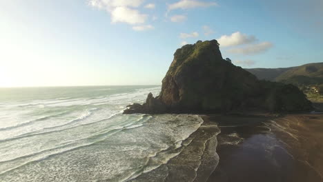 aerial wide angle of lion rock piha tracking towards as the waves gentle crash on the shore line, panning into the sunlight with a lens flare