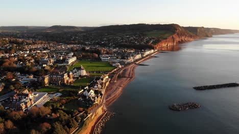 Antenne-Entlang-Der-Küste-Mit-Der-Stadt-Sidmouth,-Die-In-Goldenes-Sonnenaufgangslicht-Getaucht-Ist