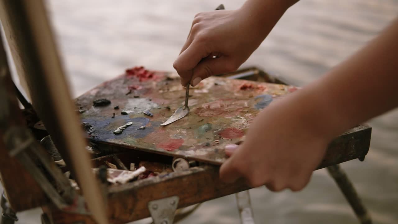 Close up shot of the women's hands, who mixes oil paints or scrape off with a palette knife, the colors are on the palette that lies on the easel. Outdoors
