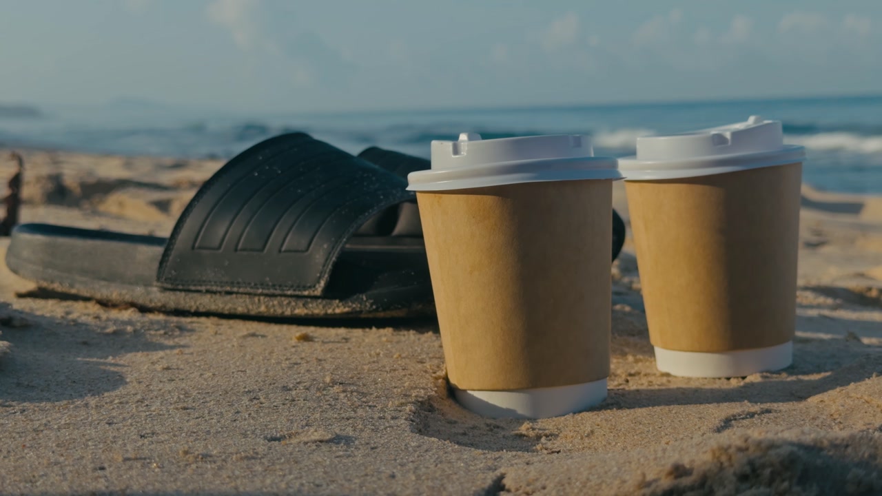 Close up of two takeaway coffee cups and a pair of slippers on a sandy beach with calm waves in the background, capturing relaxed vacation vibes and coastal lifestyle.