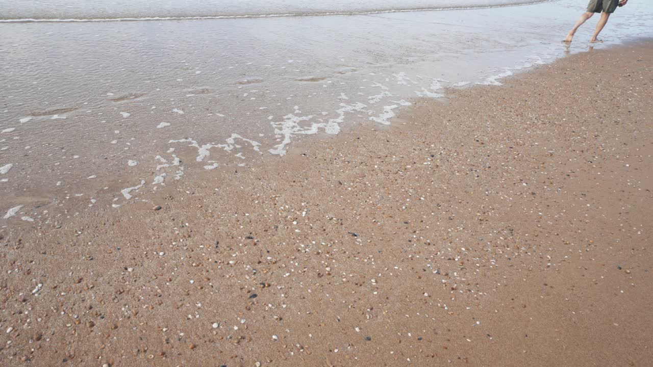 반바지를 입은 한 남자가 잔잔한 파도가 치는 해변을 따라 맨발로 모래사장을 걷고 있습니다.
