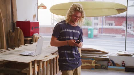 Hombre-Caucásico-Fabricante-De-Tablas-De-Surf-Parado-En-Su-Estudio-Y-Usando-Su-Teléfono-Inteligente