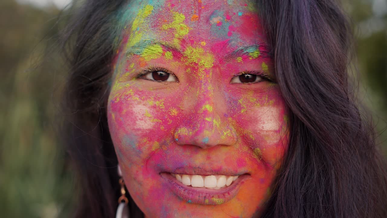 Close-up portrait of cute Asian woman with multicolored face smiling outdoors at Holi party