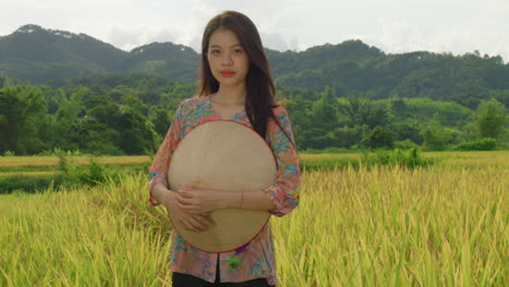 Mujer-Agricultora-Vietnamita-Parada-Frente-A-Una-Plantación-De-Arroz-Sonriendo-En-Cámara-Lenta-Vistiendo-Un-Sombrero-De-Agricultor-Con-Ropa-Tradicional