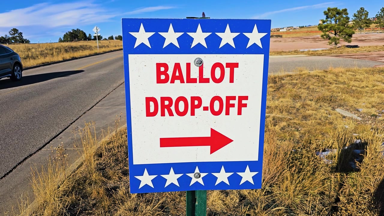 Close-up of a blue ballot drop-off sign with white stars and red lettering beside a rural road, guiding voters toward an official election ballot collection location in sunny daylight.
