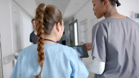 Diverse-female-doctors-discussing-work-using-tablet-walking-in-hospital-corridor,-slow-motion