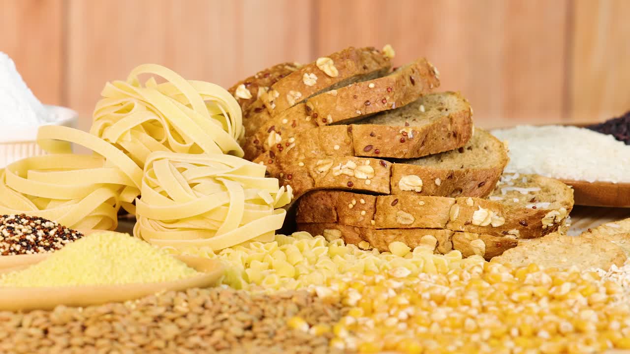 A variety of grains, pasta, and sliced bread arranged on a wooden table with warm lighting