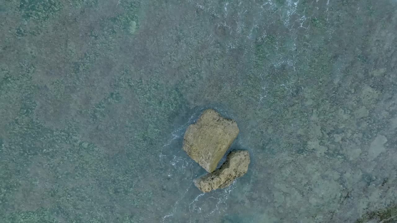 Top Down Slow Motion Drone View of Waves crashing over shallow coral reef and big rock in Uluwatu Bali Indonesia