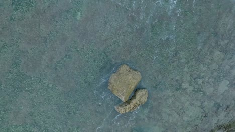 Top-Down-Slow-Motion-Drone-View-of-Waves-crashing-over-shallow-coral-reef-and-big-rock-in-Uluwatu-Bali-Indonesia