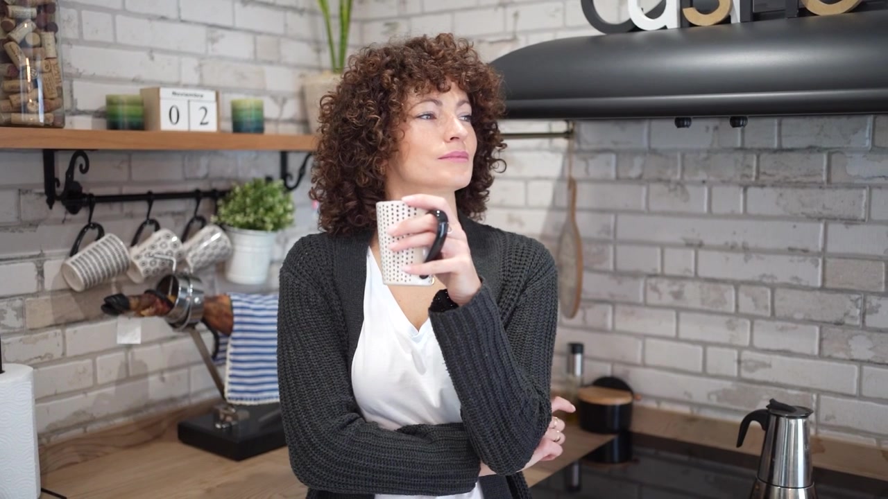 Una donna pensierosa con bellissimi capelli ricci in una cucina moderna ed elegante che tiene in mano una tazza.