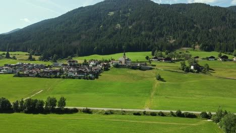 an alpine village lies nestled at the base of a forested mountain, with a prominent church and steeple at its center. lush green fields and traditional houses complete the idyllic rural scene.