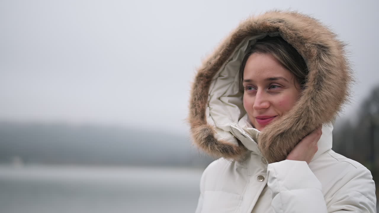 A woman stands by a calm lake, wearing a warm coat with a fur-lined hood, enjoying the peaceful winter atmosphere as she smiles gently, surrounded by nature