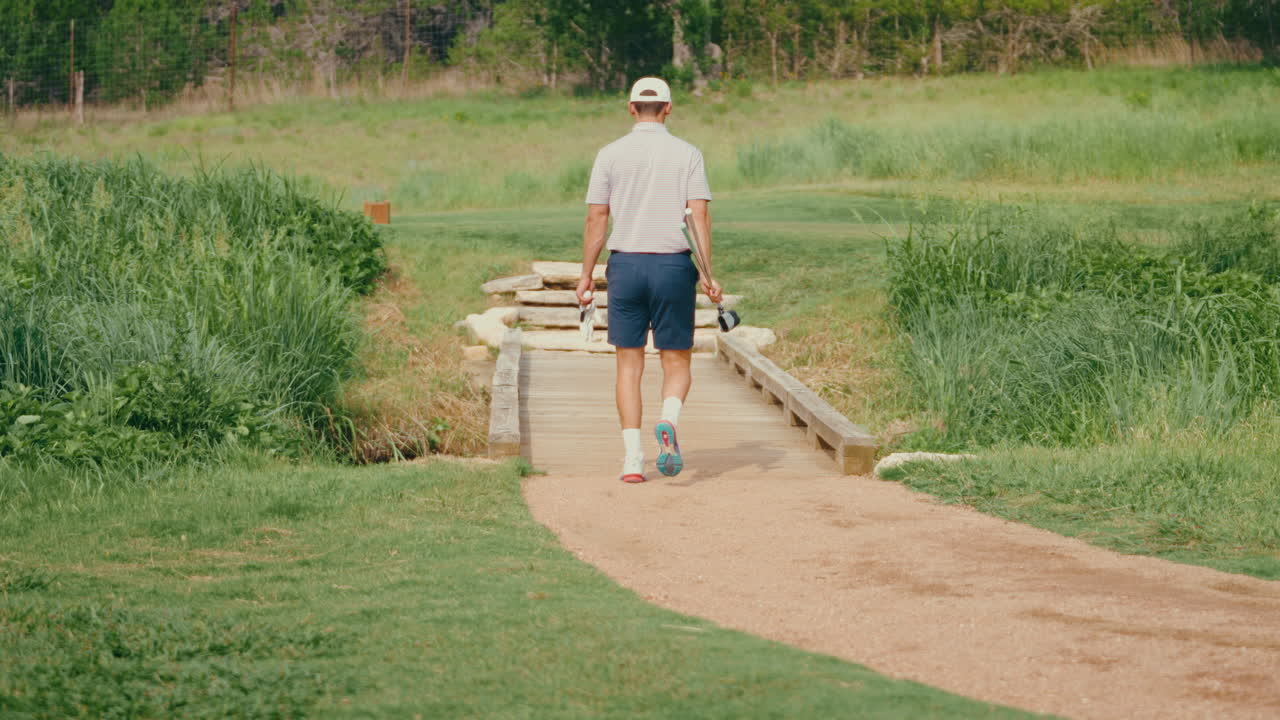 A male golfer walks away carrying multiple clubs across a scenic bridge to the tee box in slow motion. The shot captures the calm, purposeful walk and preparation moments on the golf course.