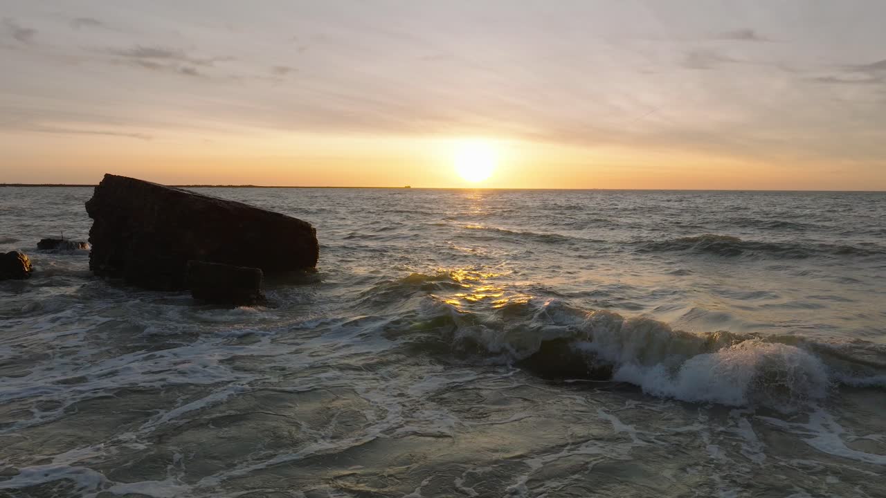 Splendida veduta aerea panoramica delle rovine delle fortificazioni costiere in cemento di Karosta, vibrante tramonto sul tempestoso Mar Baltico, sera d'inverno, ripresa drone al rallentatore che avanza a bassa quota.