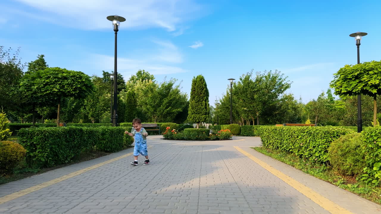 Lovely kid wearing jeans stepping happily by the paved road. Cute smiling toddler runs approaching camera. Baby walking in the park.