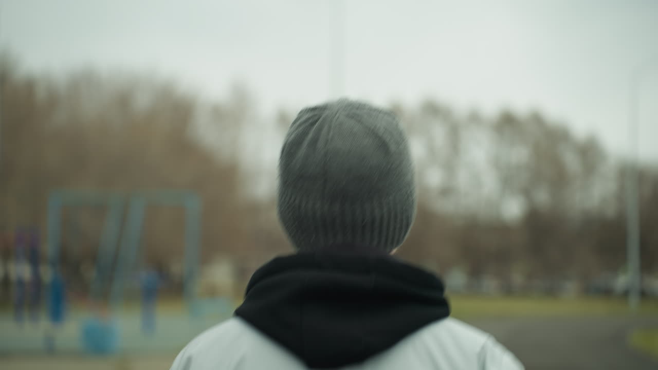 Back view of a person jogging on a track, wearing a gray jacket and a black beanie, with a blurred view of a woman in a brown jacket and another person jogging opposite each other