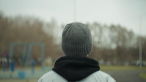 back view of a person jogging on a track, wearing a gray jacket and a black beanie, with a blurred view of a woman in a brown jacket and another person jogging opposite each other