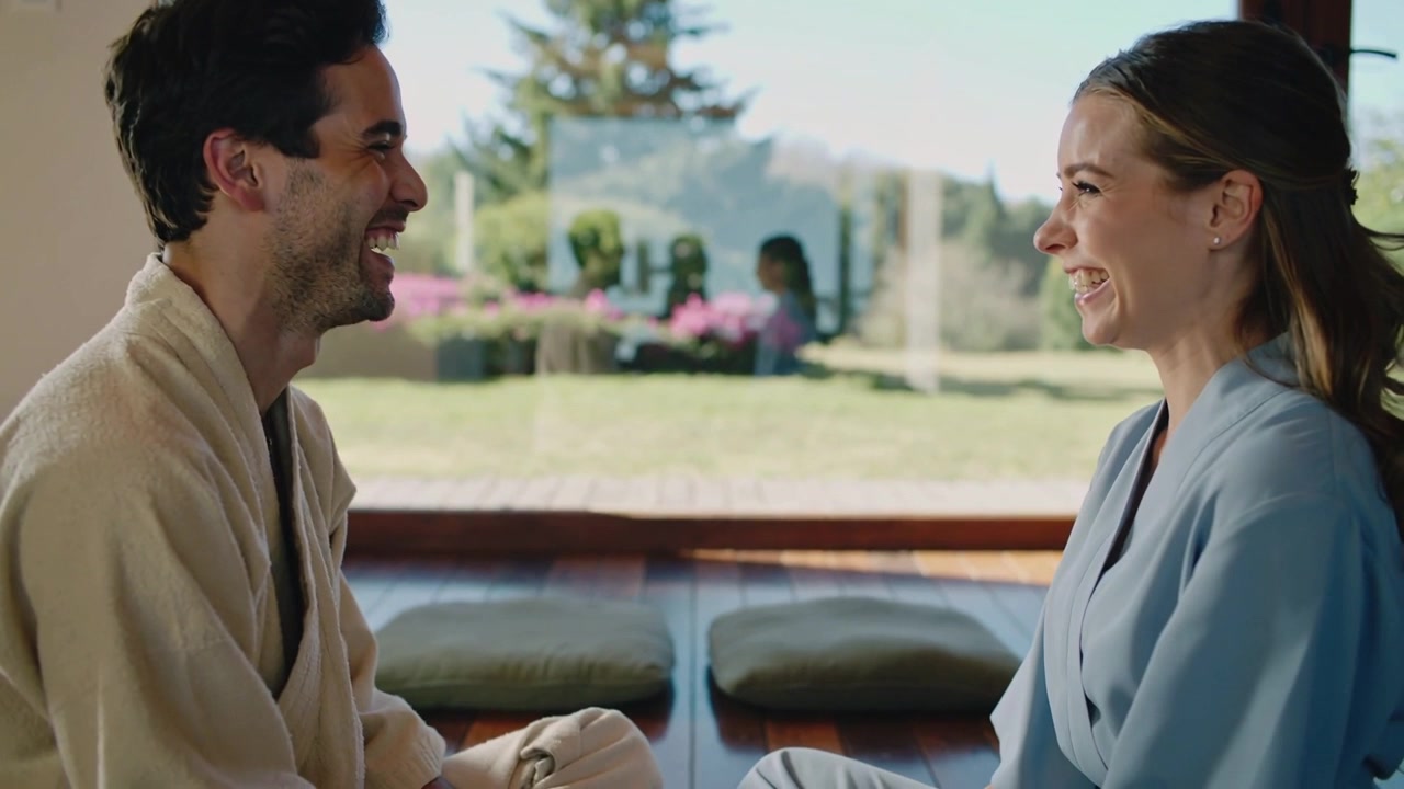 Smiling couple laughing after eye contact seated on cushions at home, robes and glass wall visible