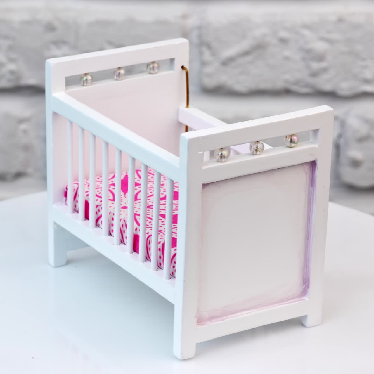 White bed for doll with decorations. Little white crib with bright pink mattress at the rotating stand. White wall background