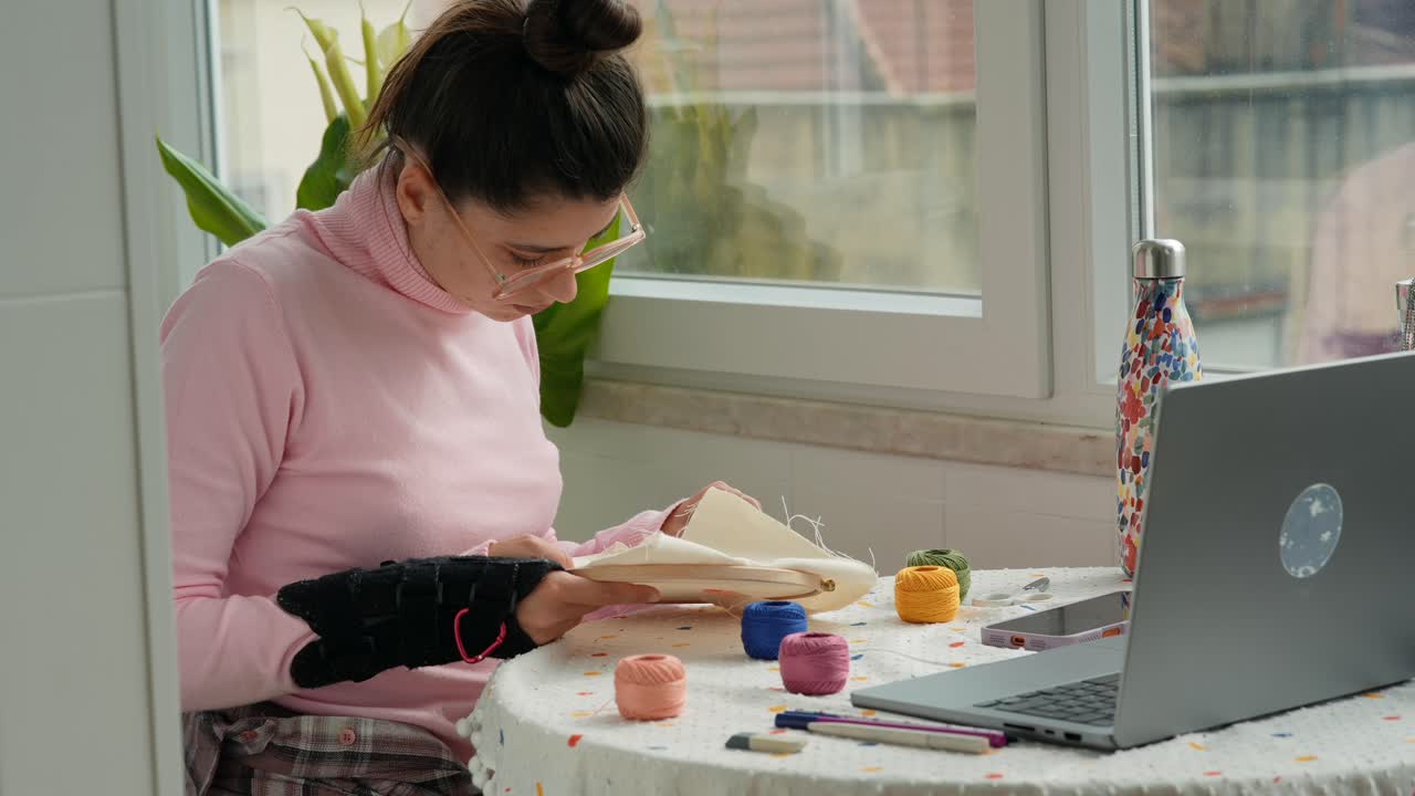 Giovane donna con tutore al polso che ricama al tavolo di casa con un laptop