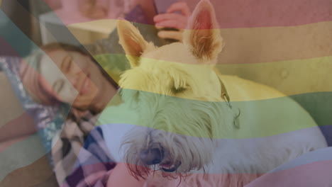 animation of pride rainbow flag over happy caucasian woman relaxing with smartphone and pet dog