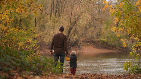 cute toddler is holding hand of his father in forest man and child are standing together on calm shore of lake at autumn day idyllic picture of famly rest at weekends