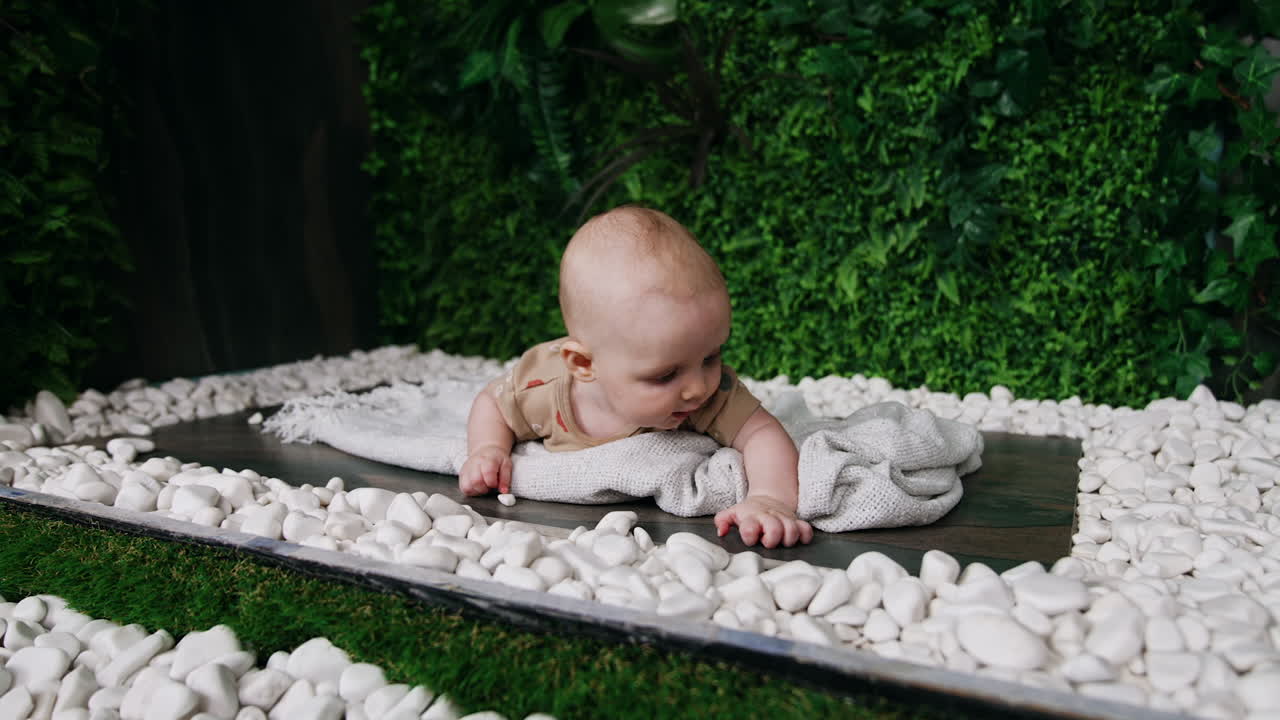Caucasian baby boy lies on the white plaid. Cute kid is interested with white pebbles around him. Artificial greenery at backdrop.