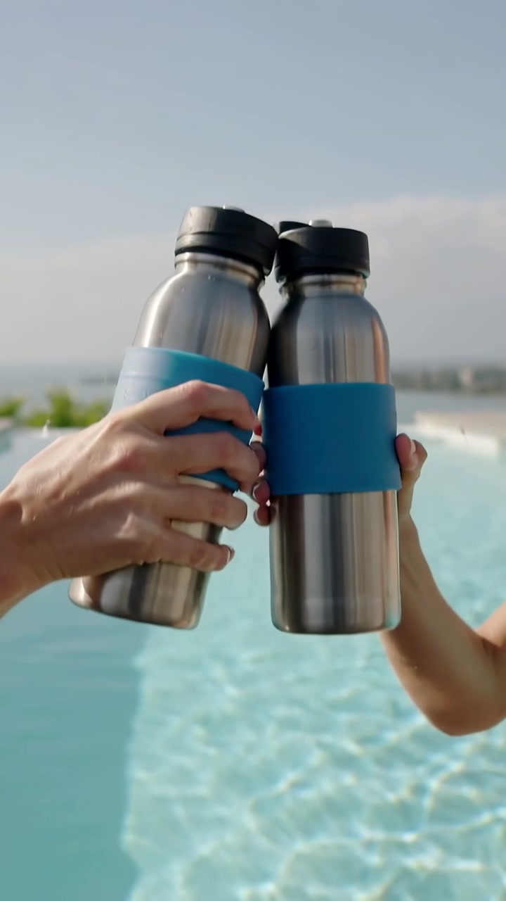 Pair clinking steel bottles with blue grips, hands bringing together, toasting at infinity pool