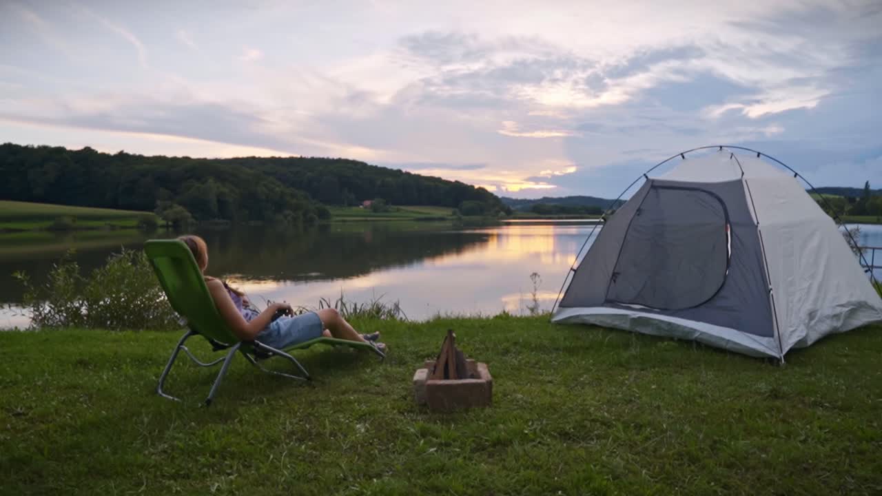 Kvinde ligger på udendørsstol nær rolig sø med telt og lejrbål ved solnedgang, nyder fredelig campingstemning og naturskønt landskab i Savci, Slovenien