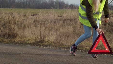 Woman-in-safety-vest-walks-to-place-traffic-safety-triangle-in-road,-pan