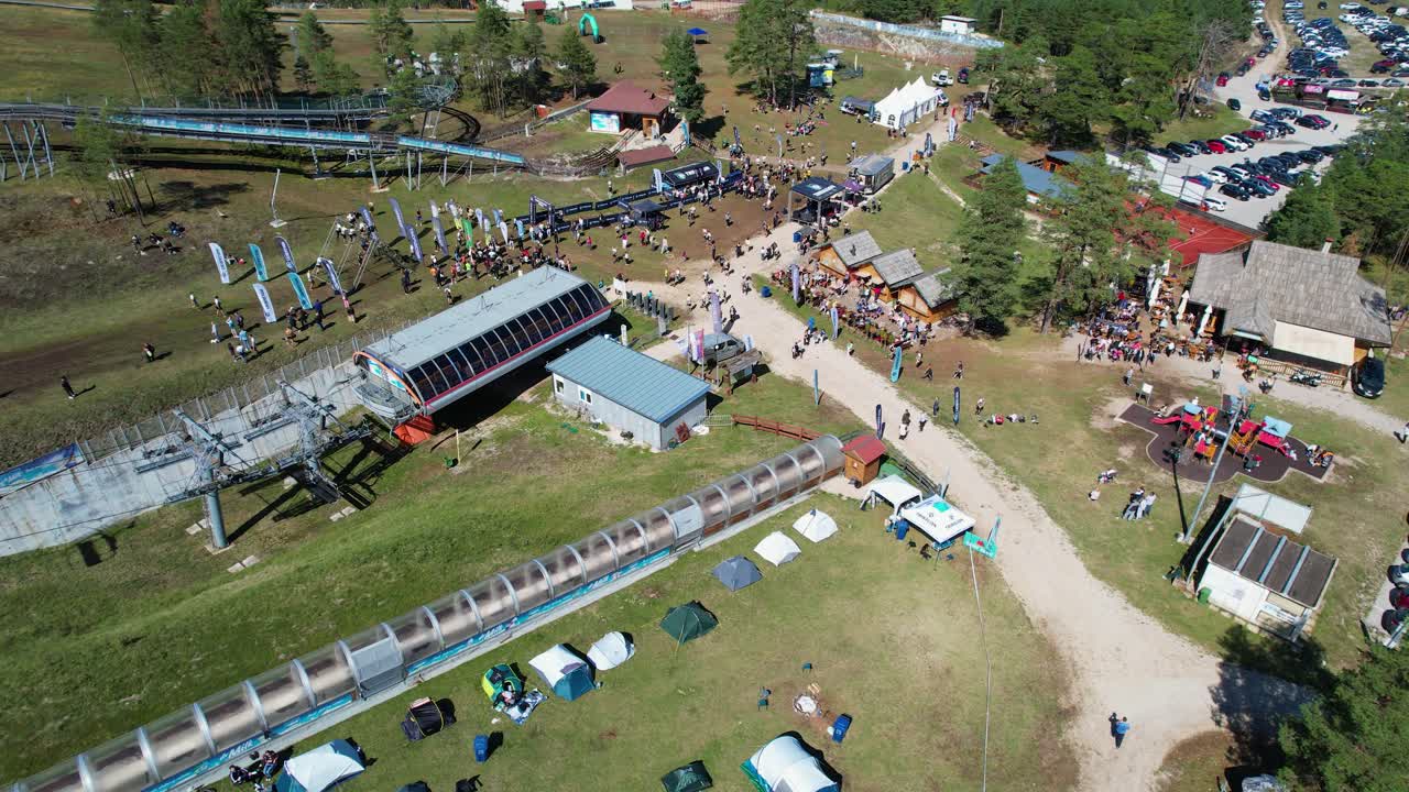 vista aérea de un animado evento al aire libre celebrado en un parque de aventuras, con tiendas de campaña, participantes, puestos de comida, un patio de recreo y un ascensor de sillas