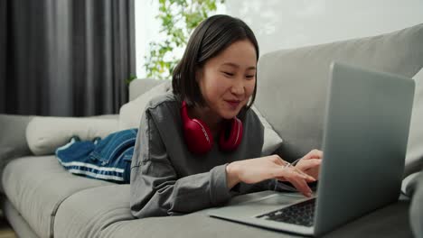 Excited-asian-woman-typing-on-laptop-while-laying-on-sofa,-communicating-with-friends