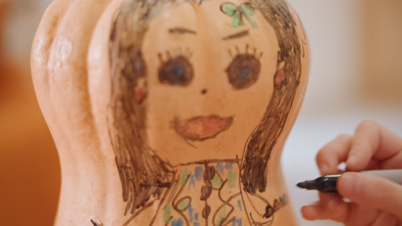Close-up pompoengezicht getekend met stift. Artiestenhand schetst een popachtig portret op kleine pompoen, voegt haar, wimpers, roze mond en lijnen voor een jurk met patroon toe. Warm binnenlicht, knutselsessie op tafel