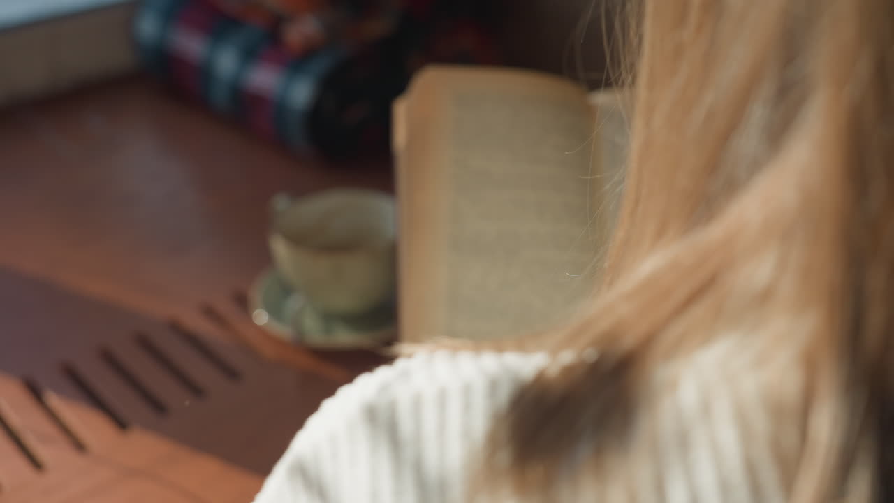 Close up back view of lady with golden hair reading book in cozy daylight as she flips pages near ceramic cup on wooden table with soft shadows cast by gentle morning sunlight