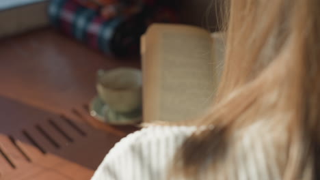 close up back view of lady with golden hair reading book in cozy daylight as she flips pages near ceramic cup on wooden table with soft shadows cast by gentle morning sunlight