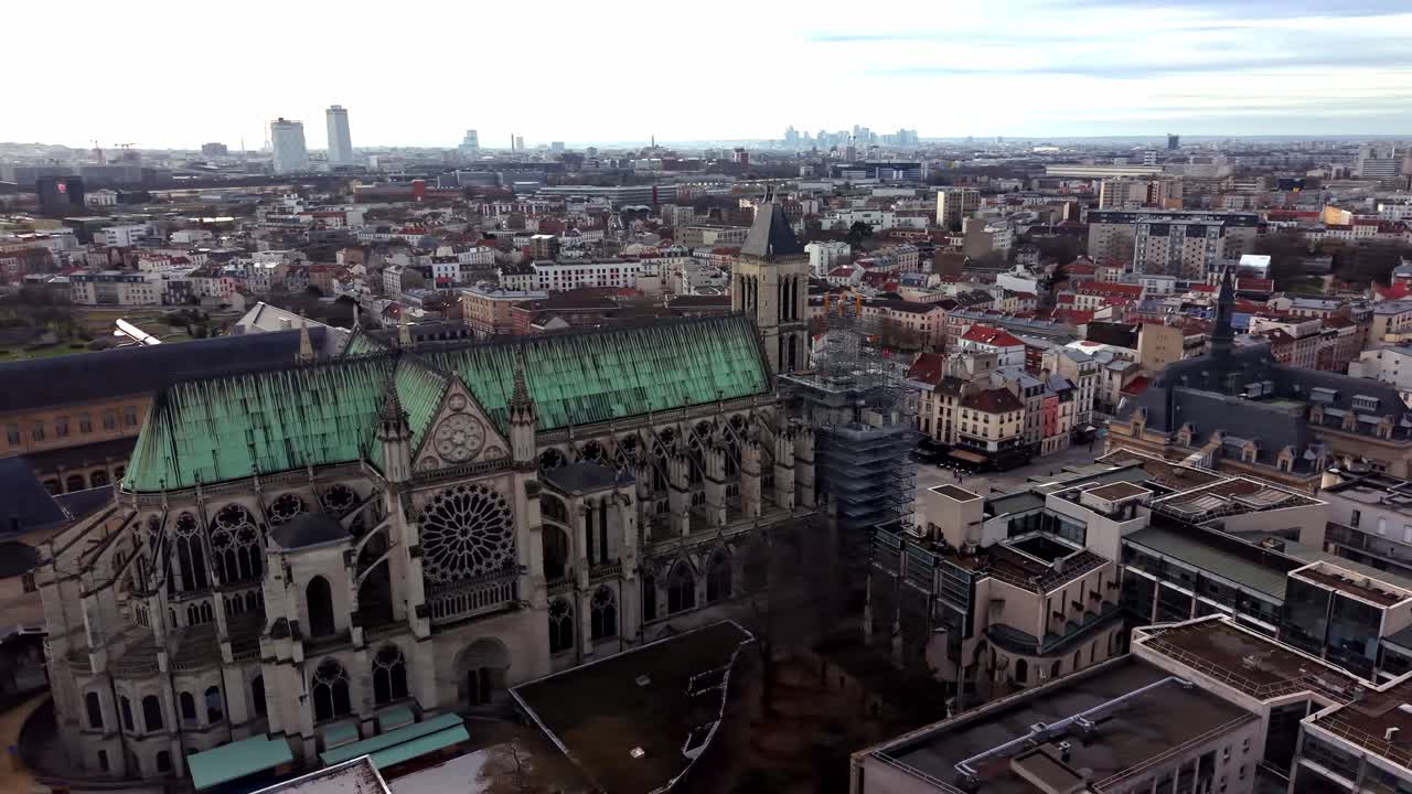 Filmisk luftfoto af den majestætiske gotiske Basilika Saint-Denis med karakteristisk grønt kobbertag og kunstfærdig arkitektur omgivet af det urbane parisiske landskab, Frankrig