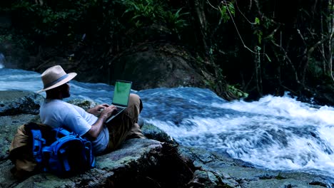 young man using green screen laptop computer on a waterfall. travel and freelance work concept