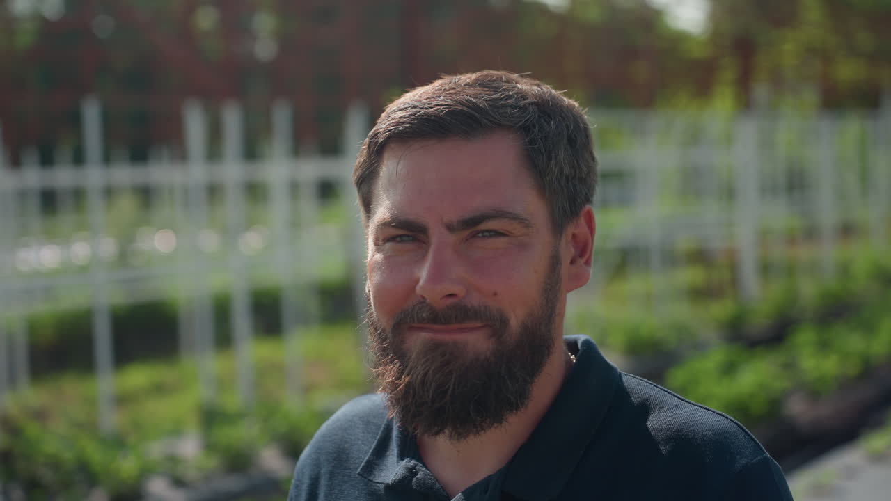 man turning and looking at camera near iron frame structure on sunny day, outdoor rural setting with green plants and blurred trees, thoughtful expression capturing connection with viewer