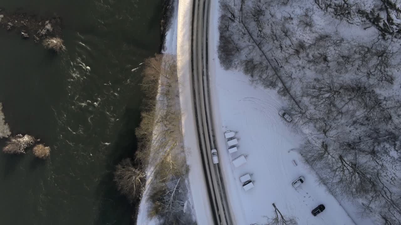 절벽과 강 사이의 구부러진 눈 덮힌 도로, 위에서 아래로 드론, 햇살