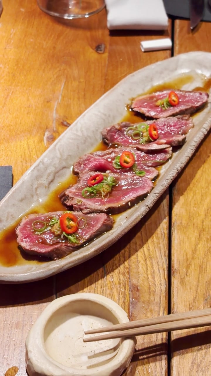 POV, tataki di manzo Wagyu raro tagliato finemente, disposto ordinatamente su un piatto di ceramica. Guarnito con anelli di peperoncino rosso e cipollotti affettati in salsa ponzu saporita su un tavolo di legno in un elegante ristorante giapponese
